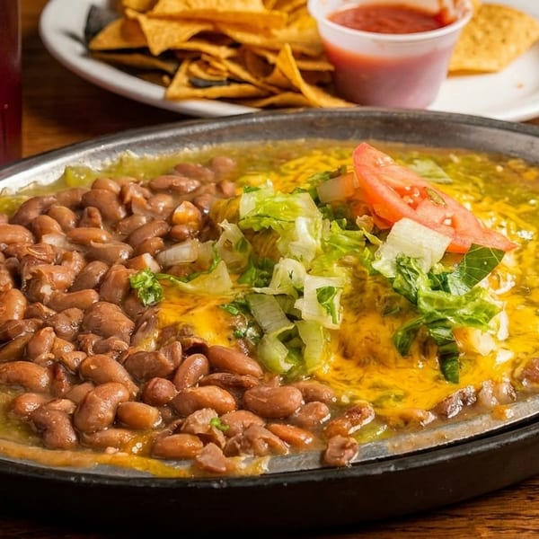 Sizzling skillet of pinto beans, green chili, melted cheese, lettuce, and tomato, with chips and salsa.
